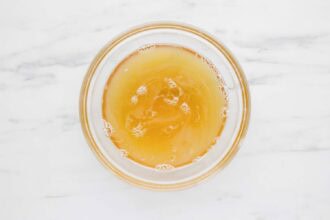 A clear glass bowl filled with light amber Mango Lemonade, viewed from above on a white marble surface.