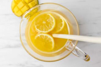 A glass pitcher filled with yellow Mango Lemonade, containing lemon slices and a white stirring stick, sits on a white surface next to a sliced mango.
