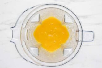 Top view of a blender containing a smooth yellow Mango Lemonade mixture on a white surface.