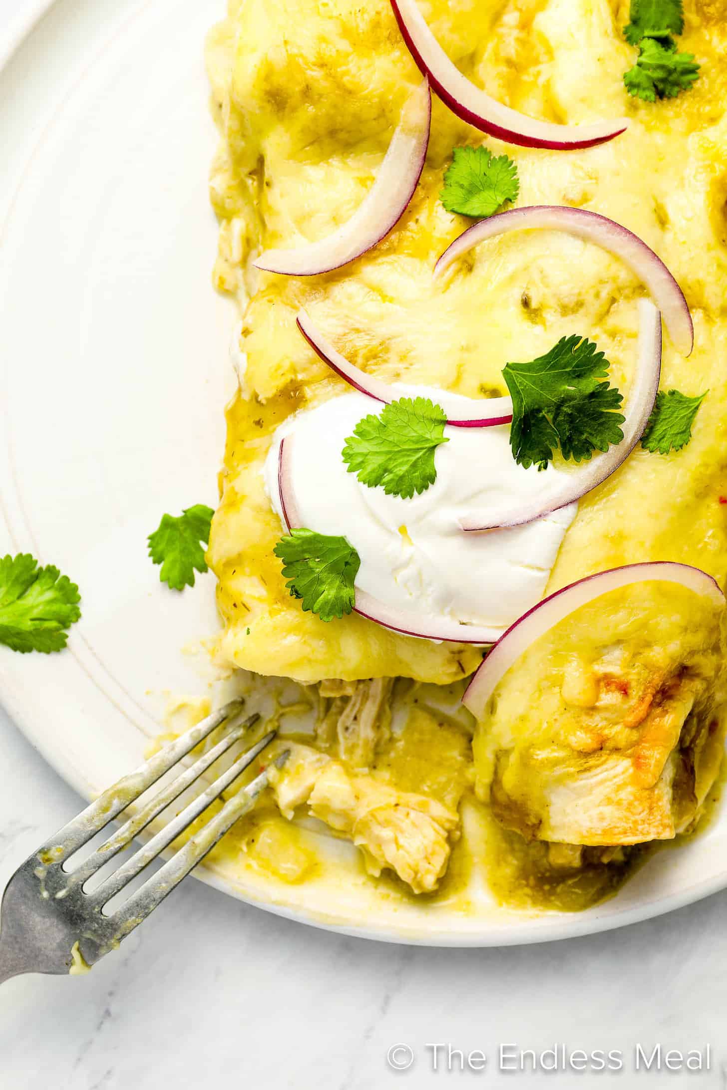 A fork taking a piece of Green Chicken Enchiladas on a dinner plate with sour cream