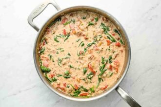 A stainless steel skillet filled with creamy sauce, ground turkey pasta, spinach, and diced tomatoes sits on a white surface.