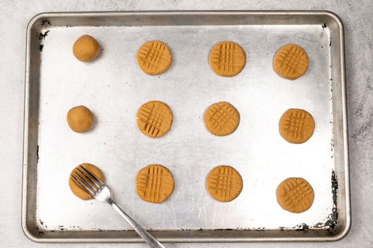 image for recipe instruction: A baking sheet with unbaked Classic Cookies—peanut butter cookies, some rolled in balls and others pressed with a fork for a crisscross pattern. A fork rests on the tray.