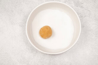 image for recipe instruction: A bowl filled with white granulated sugar and a single brown dough ball placed on top, ready to be transformed into classic cookies or delicious peanut butter cookies.