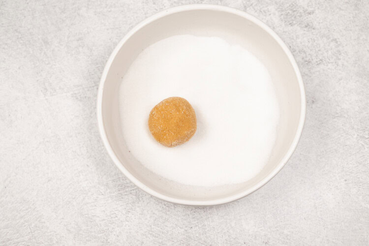 image for recipe instruction: A bowl filled with white granulated sugar and a single brown dough ball placed on top, ready to be transformed into classic cookies or delicious peanut butter cookies.