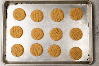 image for recipe instruction: A baking sheet with twelve evenly spaced Peanut Butter Cookies, each featuring a classic crisscross pattern on top.