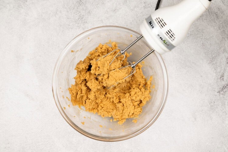 image for recipe instruction: A glass bowl containing peanut butter cookie dough is being mixed with an electric hand mixer on a light-colored surface.