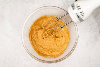 image for recipe instruction: A hand mixer is blending a light brown Peanut Butter Cookies batter in a clear glass bowl on a light-colored surface.