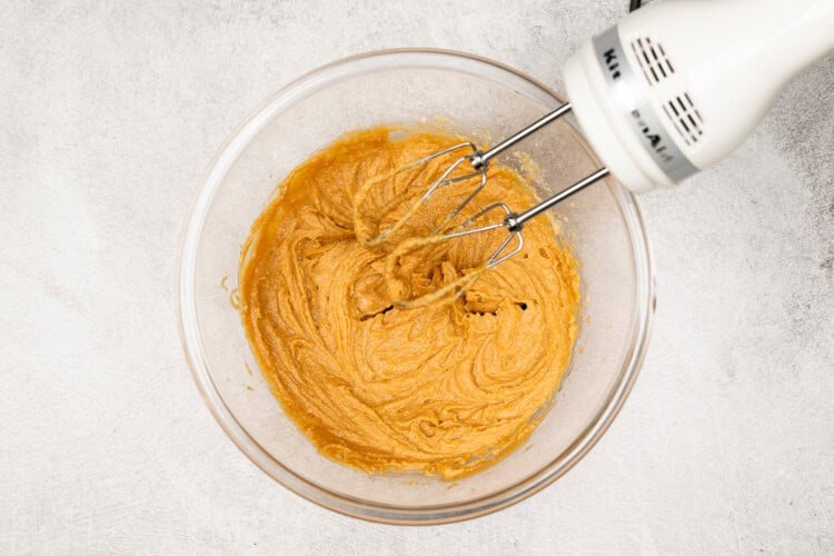 image for recipe instruction: A hand mixer is blending a light brown Peanut Butter Cookies batter in a clear glass bowl on a light-colored surface.