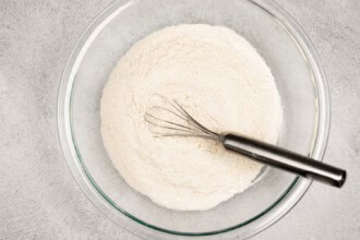 image for recipe instruction: A glass bowl containing flour with a metal whisk resting inside on a gray textured surface, ready to create classic peanut butter cookies.