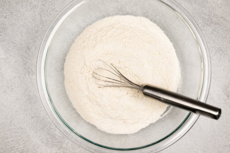 image for recipe instruction: A glass bowl containing flour with a metal whisk resting inside on a gray textured surface, ready to create classic peanut butter cookies.