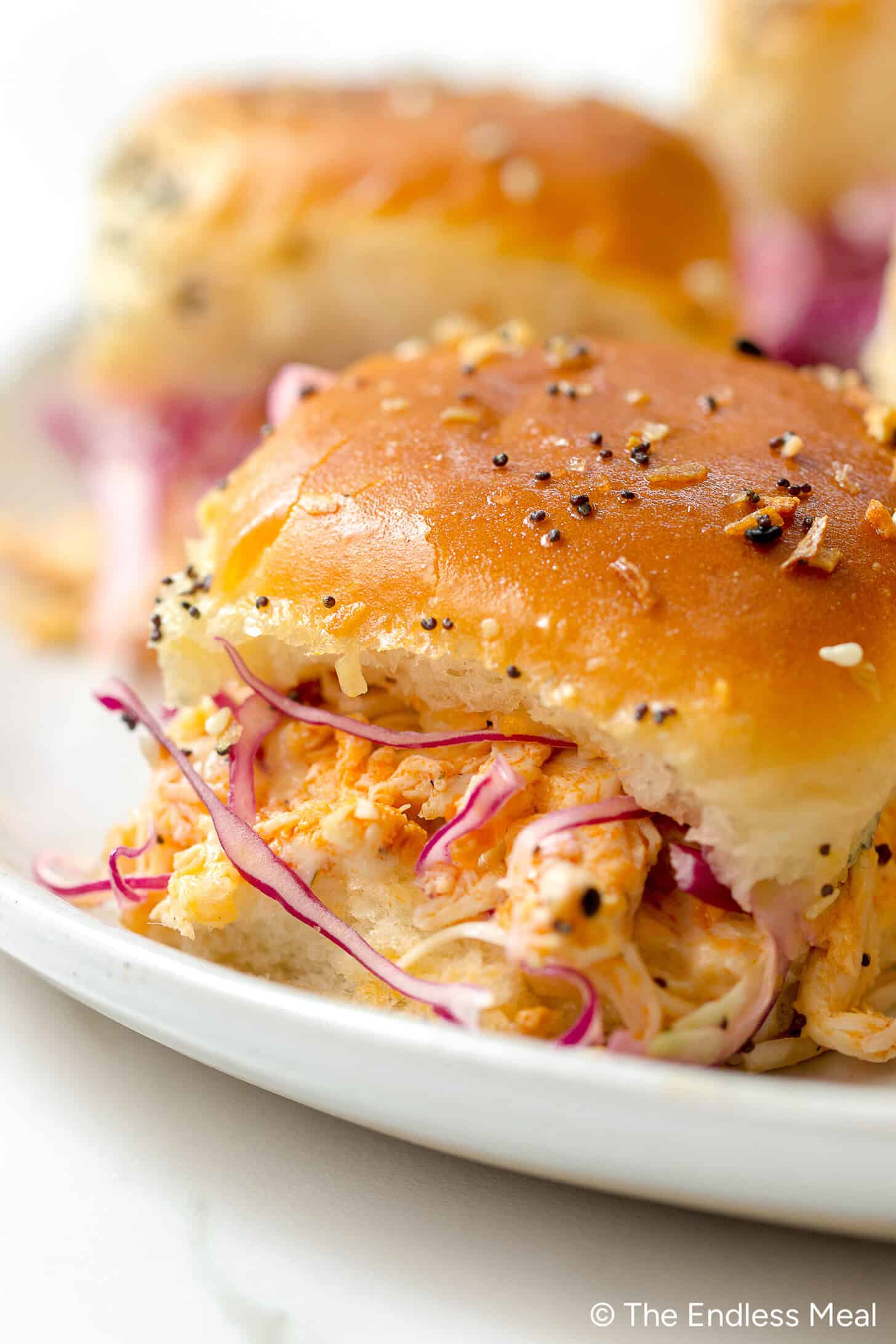 Close-up of Buffalo Chicken Sliders with tender pulled chicken and coleslaw on a white plate, topped with a golden, seeded bun.