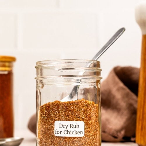 A glass jar labeled Dry Rub for Chicken, filled with flavorful chicken seasoning and a metal spoon inside, sits on a light countertop.