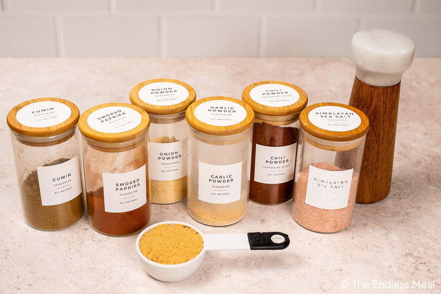 Six pots d'épices (cumin, paprika fumé, poudre d'oignon, poudre d'ail, poudre de chili, sel de l'Himalaya), un moulin à poivre et une cuillère doseuse avec de la poudre brune, parfaites pour préparer un délicieux frottement sec pour le poulet, à placer sur une surface de travail.