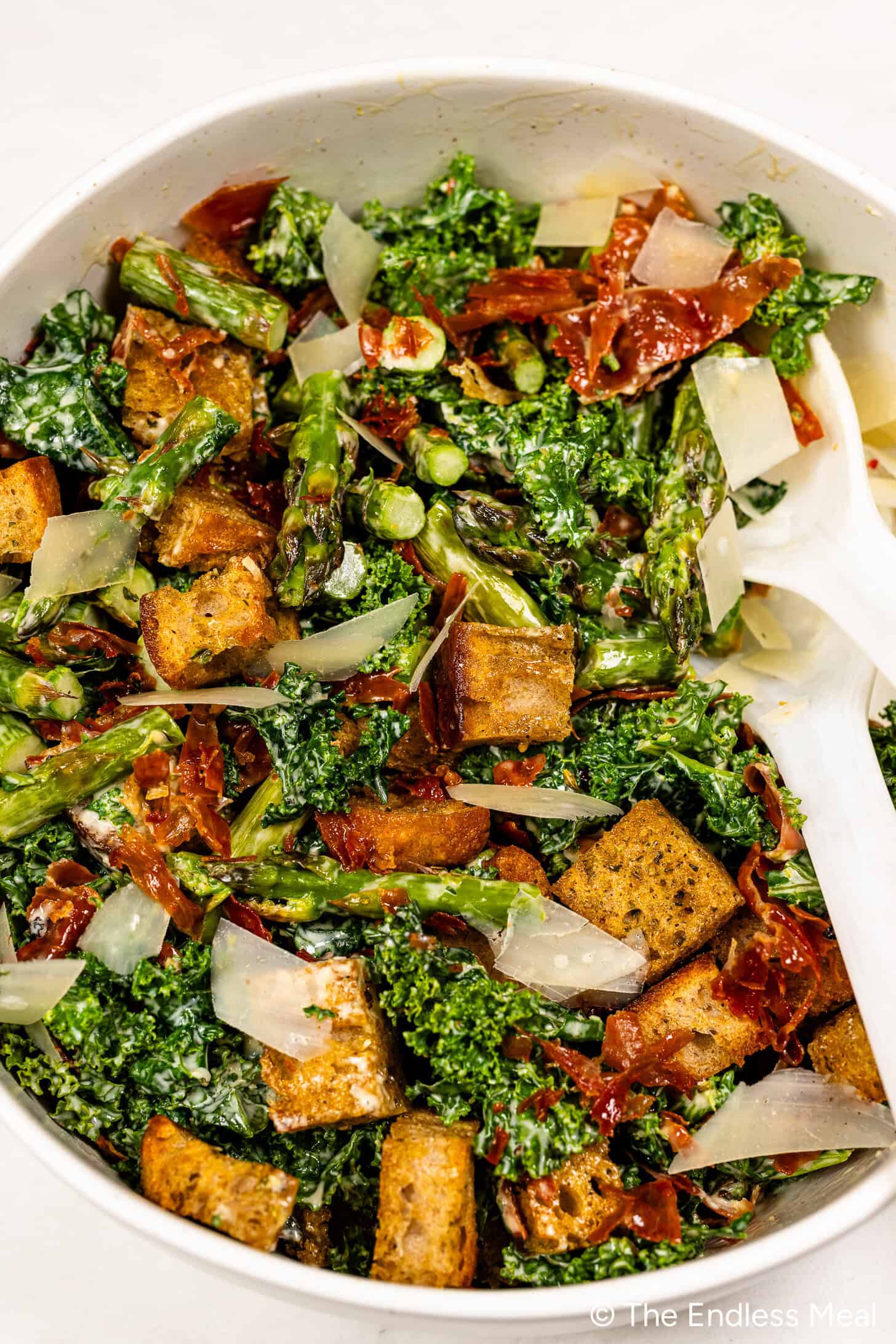 A bowl of Kale and Asparagus Caesar Salad featuring croutons, sun-dried tomatoes, Parmesan cheese, and a pair of white serving utensils.