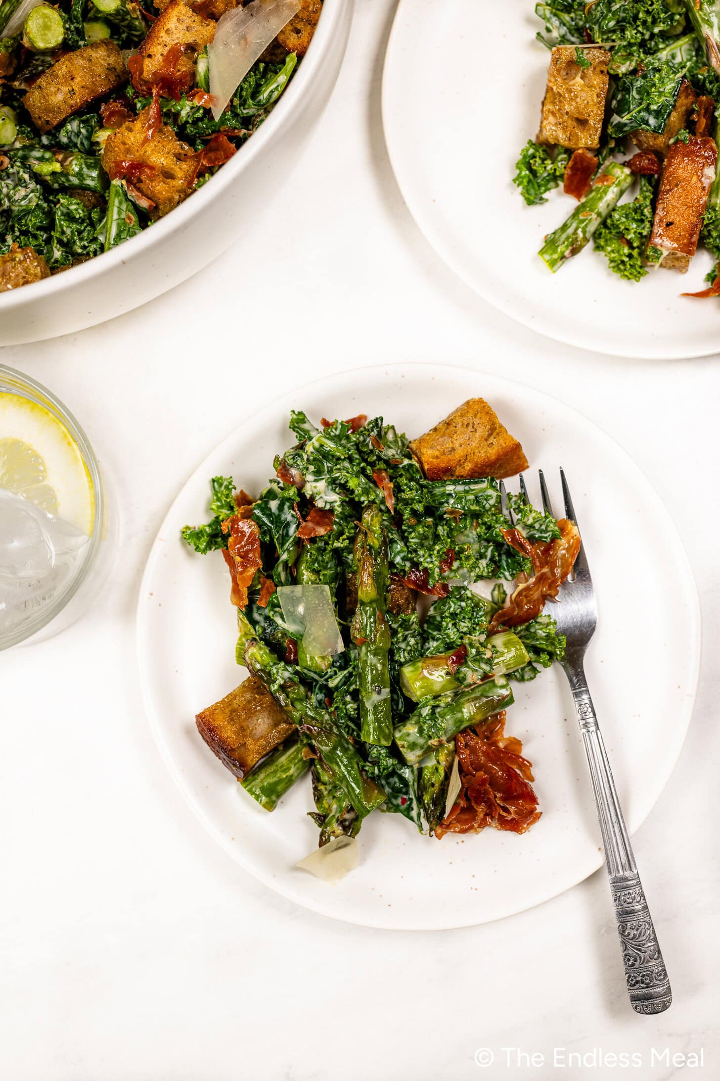A plate of Kale and Asparagus Caesar Salad with croutons, sun-dried tomatoes, parmesan shavings, and a fork beside a glass of water with lemon.