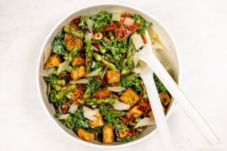 A bowl of Kale and Asparagus Caesar Salad with croutons, sun-dried tomatoes, and sliced onions, with white salad servers resting inside the bowl.