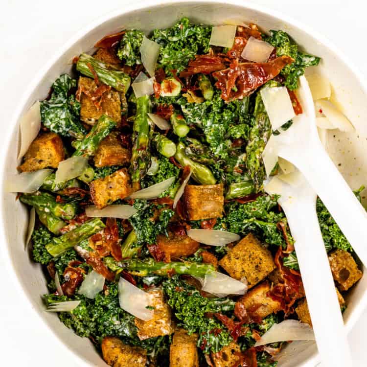 A bowl of Kale and Asparagus Caesar Salad features croutons, asparagus, and crispy prosciutto, all mixed with salad tongs.