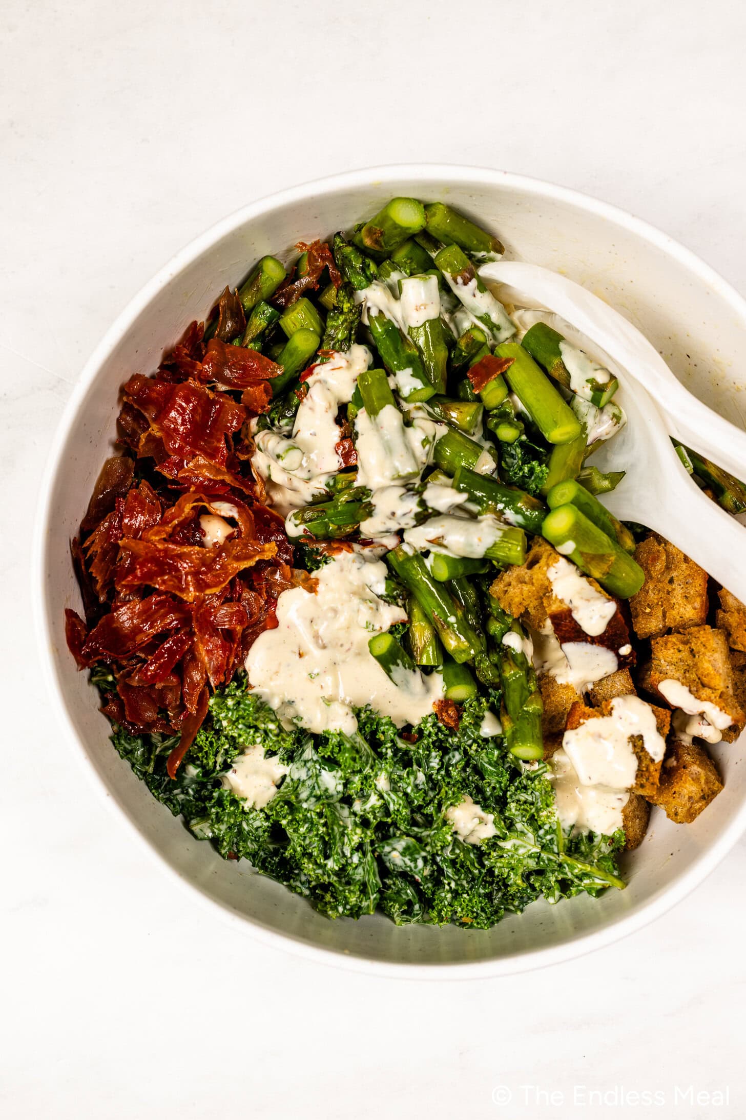 A bowl containing a Kale and Asparagus Caesar Salad with chopped kale, asparagus, crispy bread pieces, crispy prosciutto, creamy dressing, and two white serving utensils.