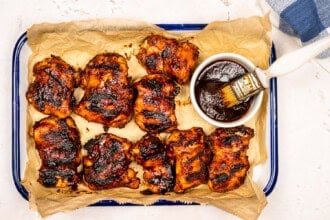 A tray of juicy grilled BBQ chicken thighs on parchment paper, served with a bowl of barbecue sauce and a basting brush.