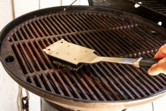 A hand uses a metal brush to clean the grates of a round, black outdoor grill, preparing it for perfectly cooked Grilled BBQ Chicken.