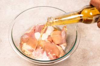 Raw chicken pieces in a glass bowl are marinated with beer, poured from a bottle, preparing them to become juicy Grilled BBQ Chicken.