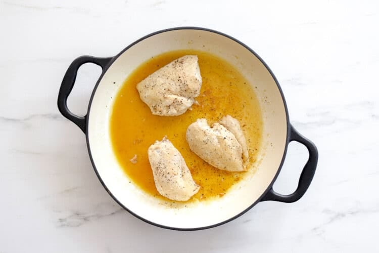 image for recipe instruction: Three partially cooked, seasoned chicken breasts in a round black-handled pan with juices, perfect for shredding to make Buffalo Chicken Sliders, placed on a white marble surface.