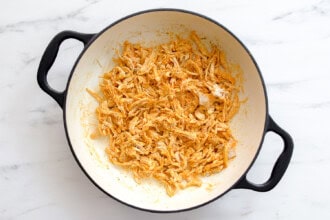 image for recipe instruction: Shredded seasoned chicken for Buffalo Chicken Sliders in a round white pot with black handles, sitting on a white marble surface.