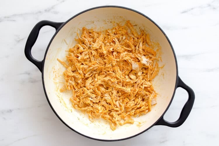image for recipe instruction: Shredded seasoned chicken for Buffalo Chicken Sliders in a round white pot with black handles, sitting on a white marble surface.