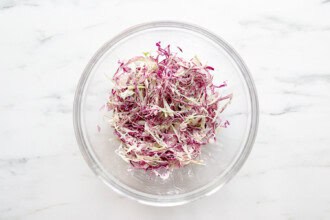 image for recipe instruction: A glass bowl containing shredded red and green cabbage, perfect for topping Buffalo Chicken Sliders, sits on a white marble surface.