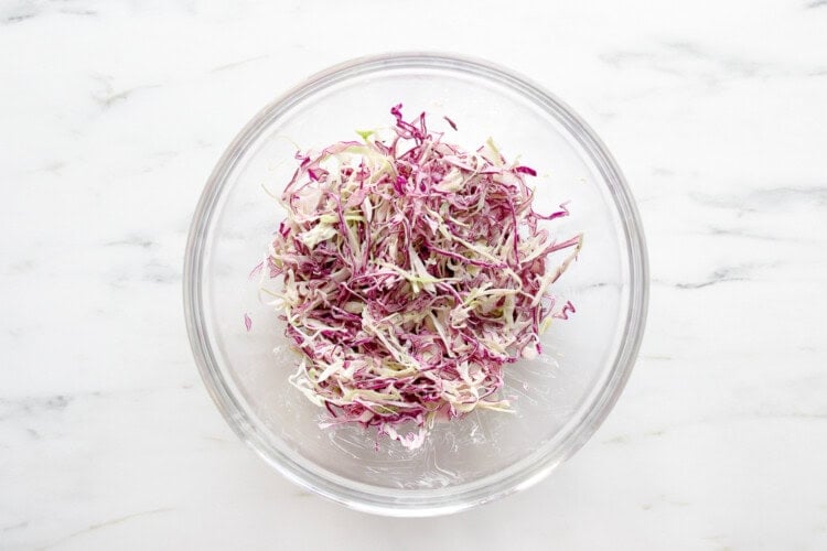 image for recipe instruction: A glass bowl containing shredded red and green cabbage, perfect for topping Buffalo Chicken Sliders, sits on a white marble surface.