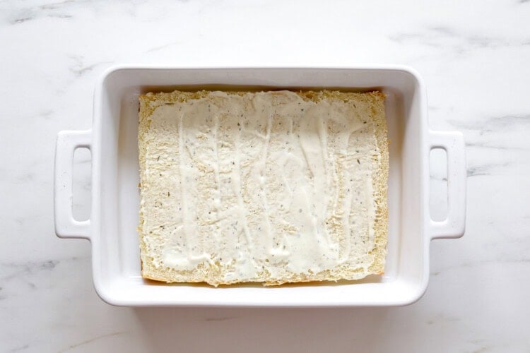 A rectangular baking dish contains a single layer of flat bread or pasta spread with a creamy white sauce, reminiscent of the base used in Buffalo Chicken Sliders, on a marble surface.