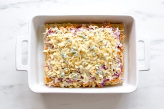 image for recipe instruction: A white rectangular baking dish filled with an uncooked shredded cheese, vegetable, and Buffalo Chicken Sliders mixture sits on a white marble surface.