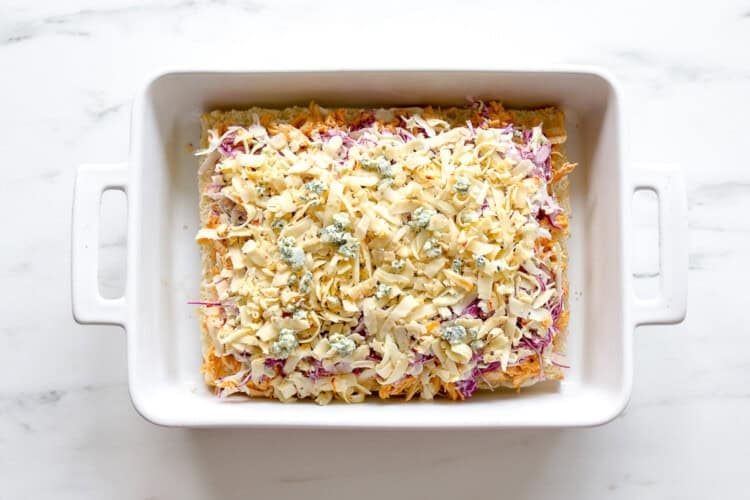 image for recipe instruction: A white rectangular baking dish filled with an uncooked shredded cheese, vegetable, and Buffalo Chicken Sliders mixture sits on a white marble surface.