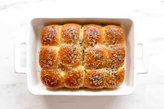 image for recipe instruction: A white baking dish contains twelve golden brown Buffalo Chicken Sliders topped with an everything bagel seasoning blend, viewed from above on a white surface.