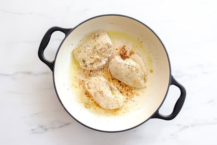 image for recipe instruction: Three seasoned chicken breasts are simmering in a white enamelled cast iron pot on a white marble surface—perfect for making flavorful Buffalo Chicken Sliders.