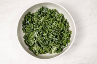 image for recipe instruction: A white bowl filled with Kale and Asparagus Caesar Salad, the chopped kale and asparagus coated in a creamy dressing, sits on a white marble surface.