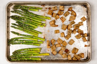 image for recipe instruction: A baking sheet with roasted asparagus spears for a Kale and Asparagus Caesar Salad on the left and seasoned croutons on the right, both arranged on a silver pan.