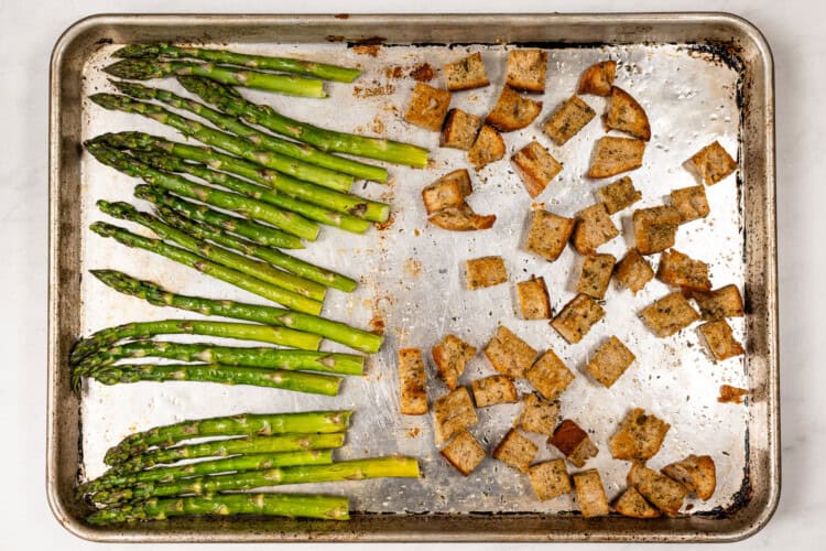 image for recipe instruction: A baking sheet with roasted asparagus spears for a Kale and Asparagus Caesar Salad on the left and seasoned croutons on the right, both arranged on a silver pan.