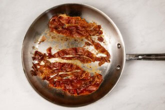 image for recipe instruction: Crispy cooked bacon strips in a stainless steel skillet on a white marble surface—perfect for topping a fresh Kale and Asparagus Caesar Salad.