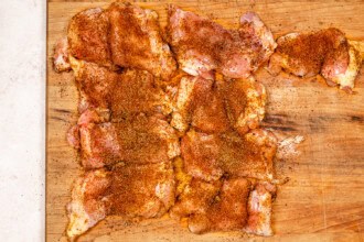 Raw chicken thighs coated with a dry spice rub are spread out on a wooden cutting board, ready to be transformed into flavorful Grilled BBQ Chicken.