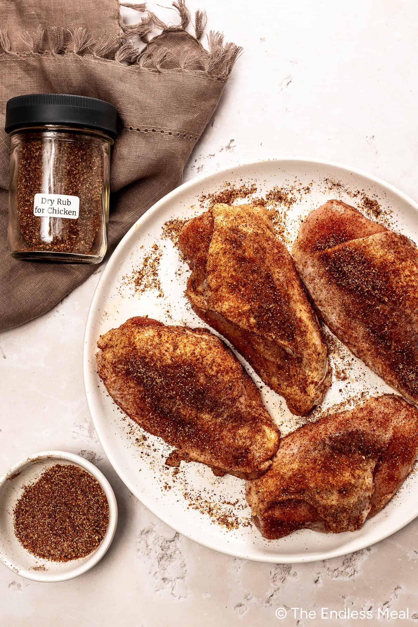 Quatre poitrines de poulet crues enrobées d'une marinade sèche d'assaisonnement de poulet placées sur une assiette blanche, accompagnées d'un pot étiqueté. 