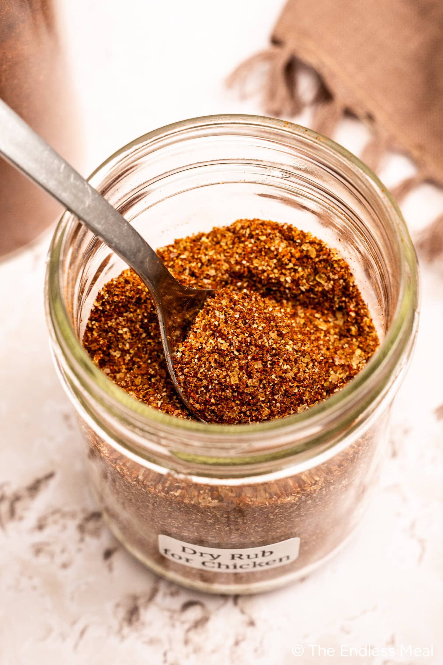 Le pot en verre étiqueté Dry Rub for Chicken contient un mélange d'épices brun rougeâtre, parfait pour assaisonner vos plats de volaille préférés, avec une cuillère en métal à l'intérieur.