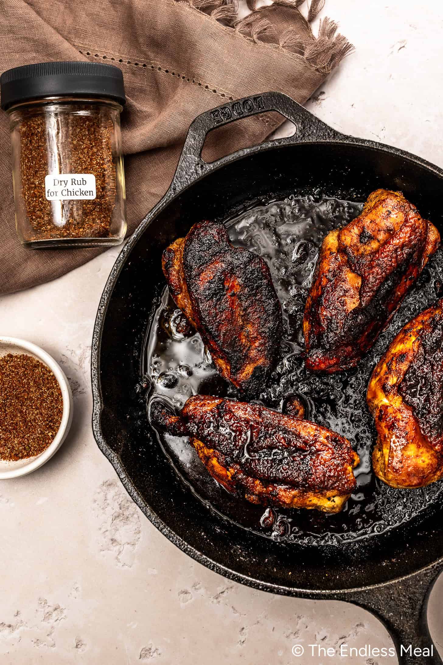 Quatre poitrines de poulet marinées et cuites dans une poêle en fonte, avec un bol étiqueté Dry Rub for Chicken et un bol du mélange d'épices signature près d'une surface de travail légère.