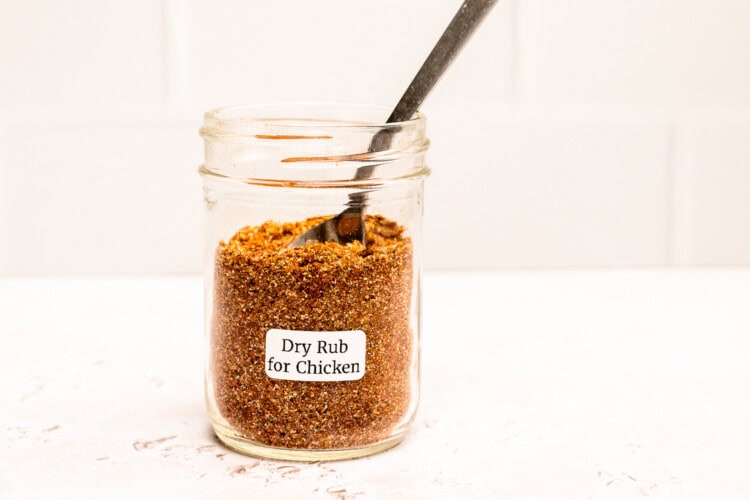 A glass jar labeled “Dry Rub for Chicken” filled with a flavorful spice mixture and a metal spoon inside, sits on a white surface, ready to add bold seasoning to your favorite chicken recipes.