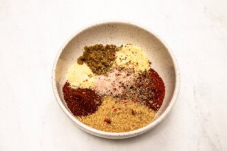 A white ceramic bowl containing separate piles of various ground spices and seasonings, perfect for creating a flavorful dry rub for chicken, sits on a white surface.