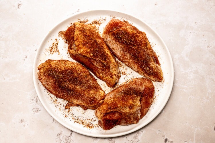 Four raw chicken breasts coated with a flavorful dry rub for chicken are arranged on a white plate atop a light-colored surface.