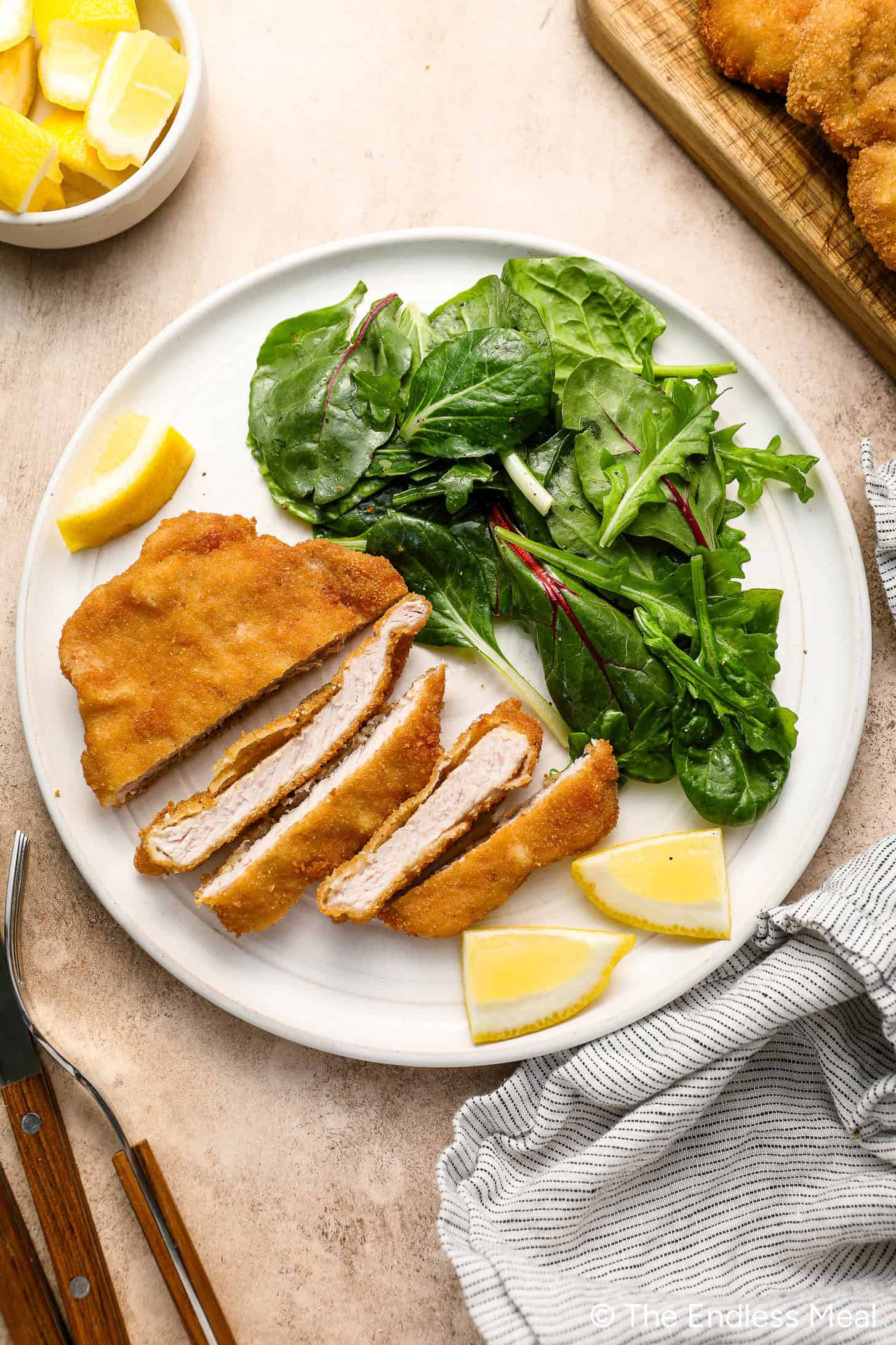 A plate with breaded and sliced crispy pork cutlet, a fresh green salad, and lemon wedges. Fork and knife are placed beside the plate.