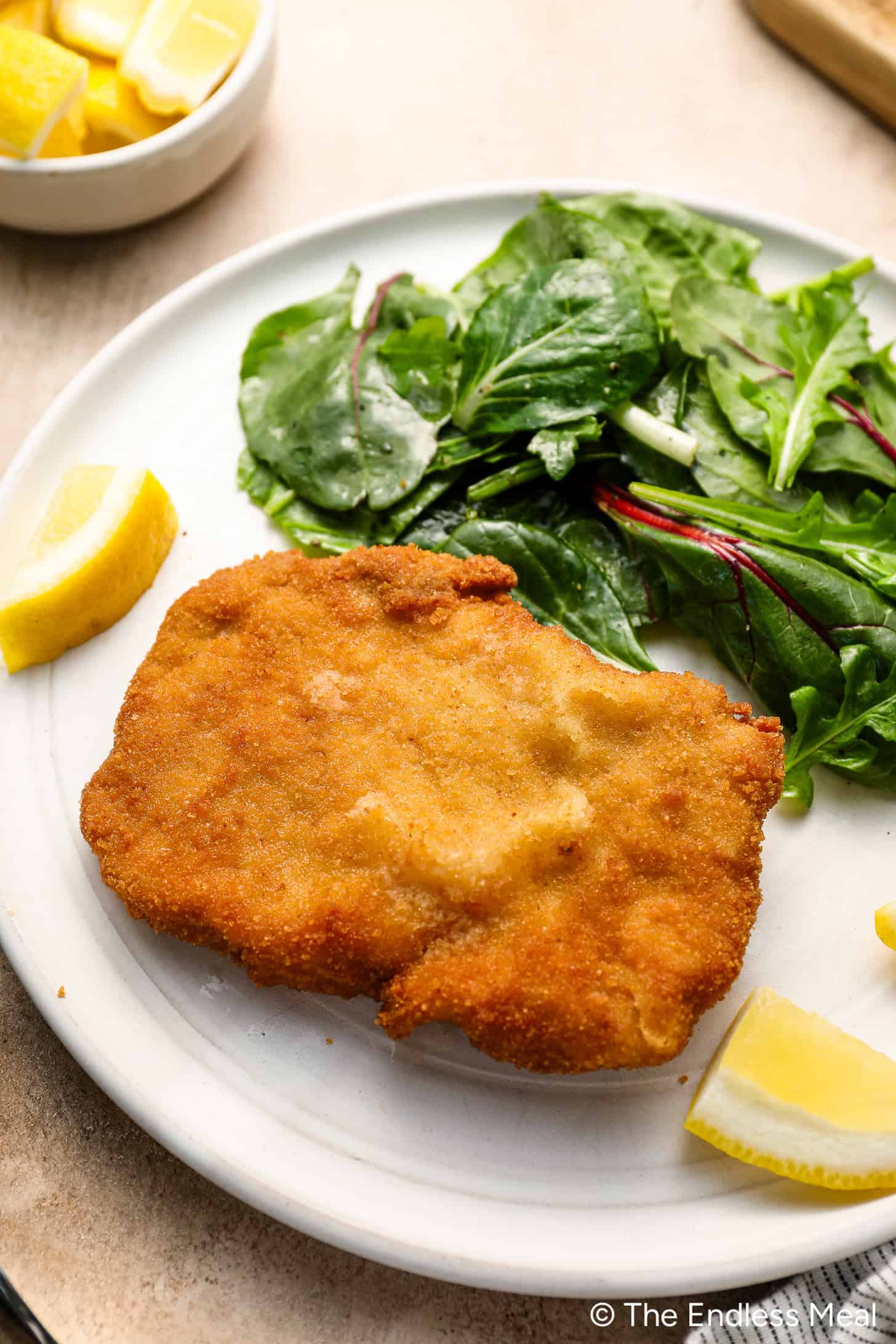 A crispy pork cutlet, breaded and fried to perfection, is served with a fresh green salad and lemon wedges on a white plate.