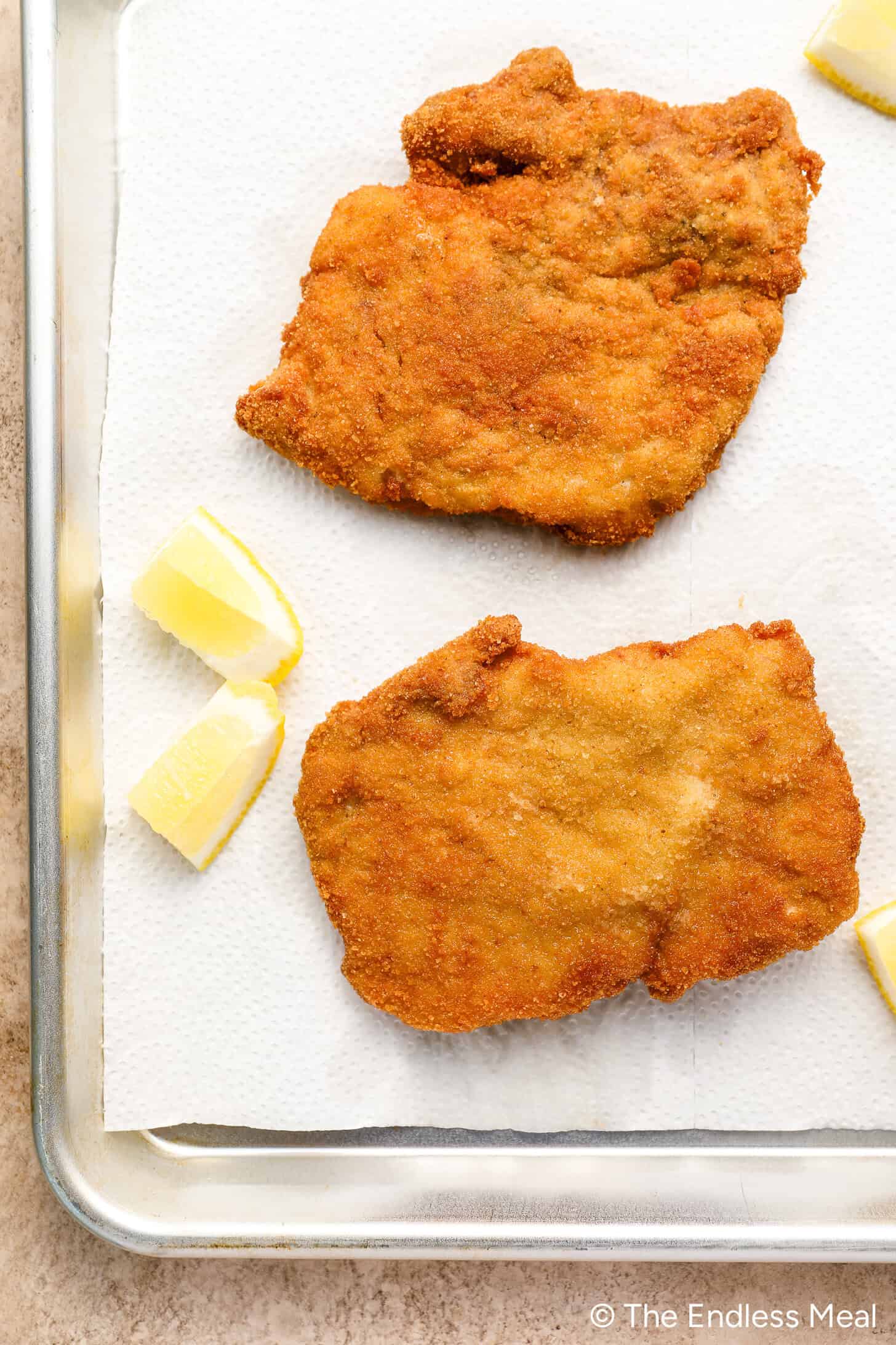 Two crispy pork cutlet schnitzel pieces rest on paper towels with three lemon wedges on a metal tray.