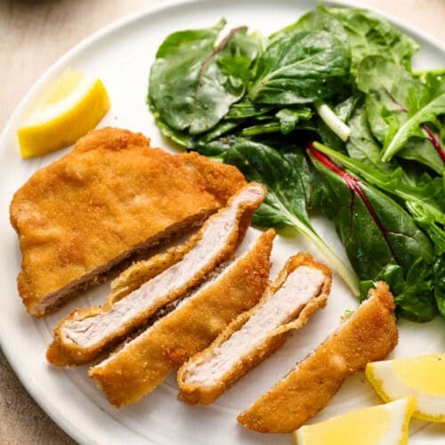 A plate with crispy pork cutlet, breaded and sliced alongside unsliced pieces, served with lemon wedges and a side of mixed green salad.